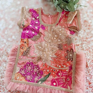 May include: A pink, embroidered dress with floral and geometric patterns. The dress features short sleeves, a round neckline, and a ruffled hem. Embellishments include sequins and gold trim.