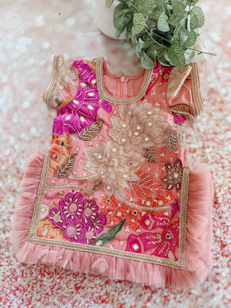 May include: A pink, embroidered dress with floral and geometric patterns. The dress features short sleeves, a round neckline, and a ruffled hem. Embellishments include sequins and gold trim.