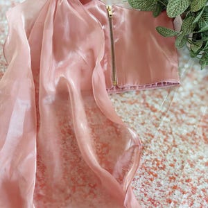 May include: A light pink satin crop top with a gold zipper and matching sheer fabric overlay. The top has a square neckline and thin straps. The sheer fabric drapes over the shoulders. The garment is displayed on a textured surface.