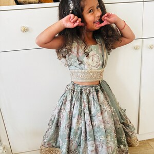 May include: A young girl wearing a light green floral print dress with gold trim and a matching top. She is smiling and has a flower crown in her hair.