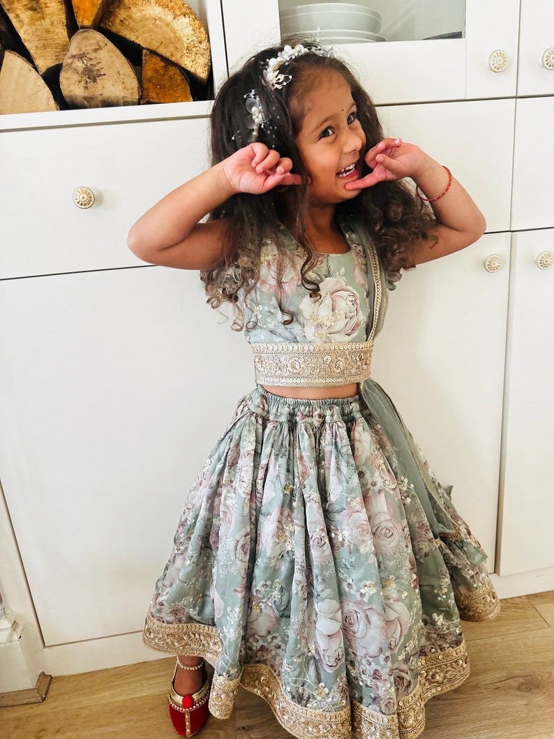 May include: A young girl wearing a light green floral print dress with gold trim and a matching top. She is smiling and has a flower crown in her hair.