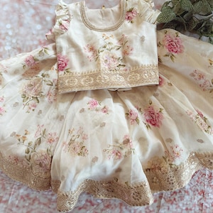 May include: A cream-colored floral two-piece outfit featuring a top and skirt. The top has ruffled sleeves and a beaded neckline. The skirt and top are adorned with pink floral patterns and gold sequin trim.