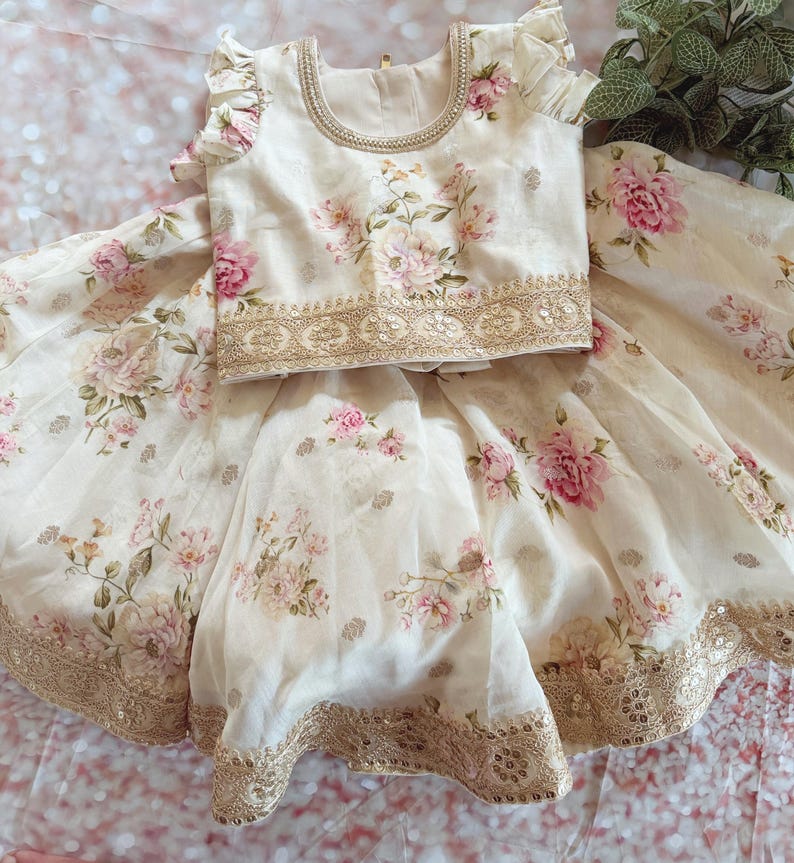 May include: A cream-colored floral two-piece outfit featuring a top and skirt. The top has ruffled sleeves and a beaded neckline. The skirt and top are adorned with pink floral patterns and gold sequin trim.