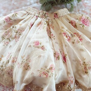 May include: A cream-colored skirt with a floral pattern of pink roses and green leaves. The skirt has a gold sequined trim along the hemline and a gathered waistband. The fabric appears to be lightweight and flowing.