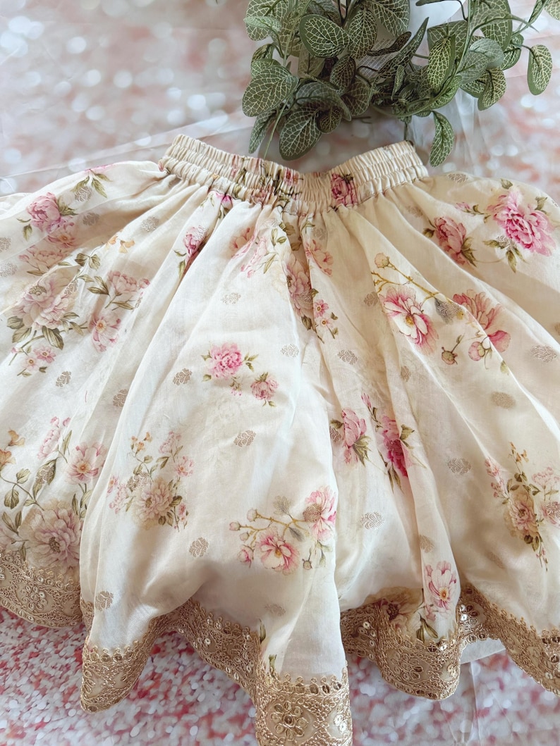 May include: A cream-colored skirt with a floral pattern of pink roses and green leaves. The skirt has a gold sequined trim along the hemline and a gathered waistband. The fabric appears to be lightweight and flowing.