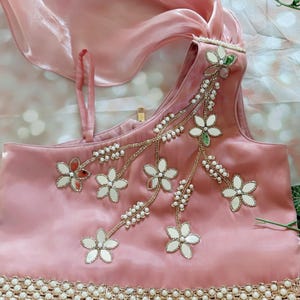 May include: A light pink satin top with floral embroidery and mirror accents. The top features a one-shoulder design and is adorned with pearls and gold trim. A matching pink scarf is draped over the top.