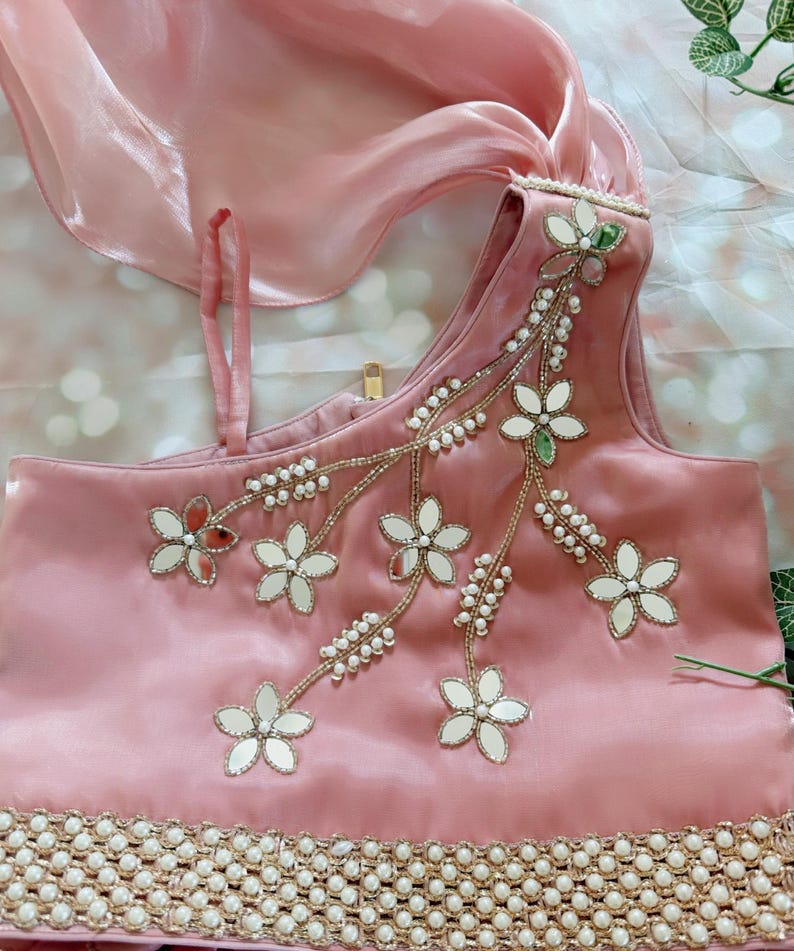 May include: A light pink satin top with floral embroidery and mirror accents. The top features a one-shoulder design and is adorned with pearls and gold trim. A matching pink scarf is draped over the top.