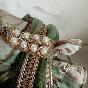 May include: A green and brown fabric with gold trim and a pearl and rhinestone embellishment.