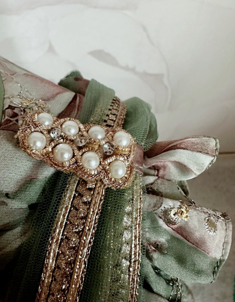 May include: A green and brown fabric with gold trim and a pearl and rhinestone embellishment.