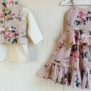 May include: A matching set of children's clothing featuring a floral print with pink roses. The girl's dress has a ruffled skirt and a gold trim. The boy's outfit includes a white shirt, a vest with a floral print, and pants. The text "Orhni bhai" and "little orhni" is visible on the clothing.