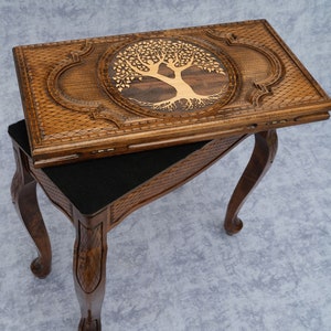 Puede incluir: Una mesa de juego de madera con un diseño de árbol de la vida tallado en la parte superior. La mesa tiene un forro de fieltro negro y cuatro patas.