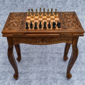 Puede incluir: Una mesa de ajedrez de madera con intrincados tallados en la parte superior y las patas. La mesa tiene una tapa abatible que revela un tablero de ajedrez con casillas negras y blancas. El tablero de ajedrez está configurado con piezas de ajedrez negras.