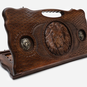 Puede incluir: Un juego de backgammon de madera con un diseño de león tallado en la tapa. La tapa está abierta y el tablero de juego es visible. El tablero de juego está hecho de madera y tiene un acabado marrón oscuro.