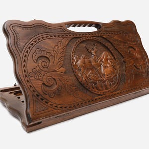 May include: A wooden backgammon board with intricate carvings. The dark brown board features a central oval relief of a deer in a mountain landscape. Decorative floral and geometric patterns frame the central image and edges.