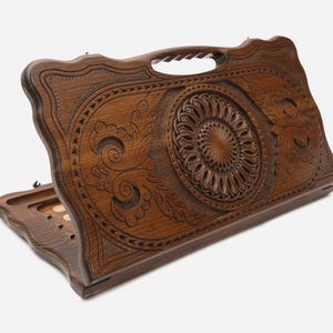 Puede incluir: Un juego de backgammon de madera con intrincados diseños florales y geométricos tallados. La madera marrón oscuro presenta un patrón circular central y un asa. El tablero de juego está abierto, revelando la superficie de juego.