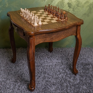 May include: A wooden chess table with a brown finish and intricate carvings. The table has a chessboard inlay and comes with a set of chess pieces.