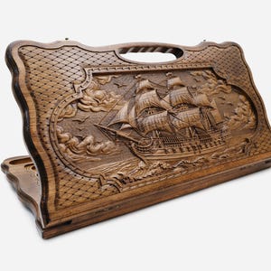 May include: A dark brown wooden backgammon board featuring an intricate relief carving of a large sailing ship on the exterior lid. The design includes detailed waves, clouds, and a cross-hatched border pattern with a built-in carrying handle.