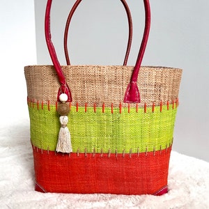 Peut inclure: Un grand sac fourre-tout tissé avec des anses en cuir rouge. Le sac est fait de fibres naturelles et présente trois bandes horizontales de couleur marron, verte et orange. Une petite perle en bois avec une perle blanche et un pompon pendent de la poignée.