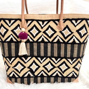 Peut inclure: Sac fourre-tout tissé avec un motif géométrique noir, blanc et beige. Le sac a des poignées en cuir et un pompon décoratif avec un pompon violet. Le sac est conçu avec une forme structurée et un fond plat.