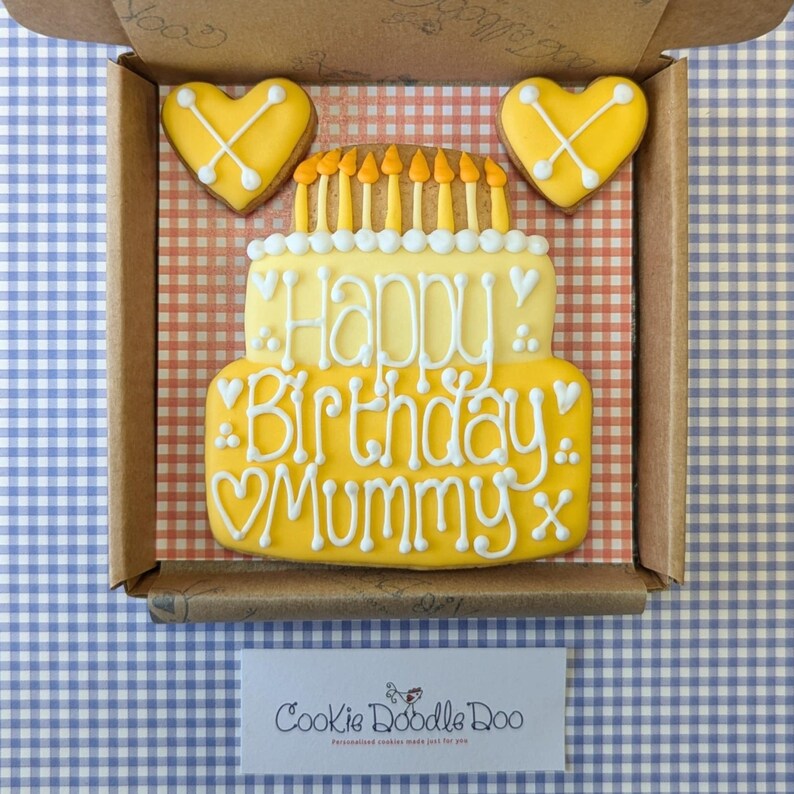 May include: A yellow and white decorated cookie in the shape of a birthday cake with the text "Happy Birthday Mummy" and two yellow heart-shaped cookies with an X design.