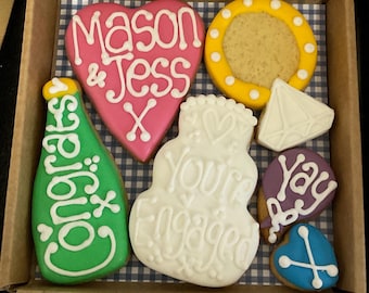 Peut inclure: Une boîte de biscuits décorés pour une fête de fiançailles. Les biscuits sont en forme de cœur, de bouteille de champagne, de gâteau de mariage, de bague de fiançailles, de ballon et de cœur avec un X. Le biscuit en forme de cœur est rose avec un glaçage blanc et dit "Mason & Jess X". Le biscuit en forme de bouteille de champagne est vert avec un glaçage blanc et dit "Félicitations". Le biscuit en forme de gâteau de mariage est blanc avec un glaçage blanc et dit "Vous êtes fiancés". Le biscuit en forme de bague de fiançailles est blanc avec un glaçage blanc. Le biscuit en forme de ballon est violet avec un glaçage blanc et dit "Youpi !". Le biscuit en forme de cœur avec un X est bleu avec un glaçage blanc.