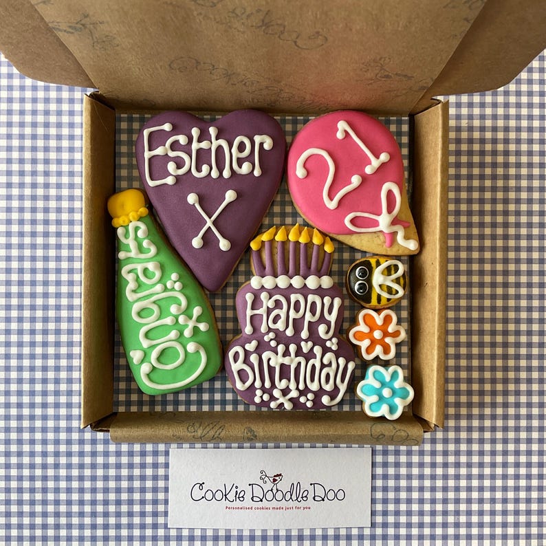May include: A box of decorated cookies with the text "Esther X" on a purple heart-shaped cookie, "21" on a pink balloon-shaped cookie, "Happy Birthday" on a purple cake-shaped cookie, and "Congrats" on a green bottle-shaped cookie. The box also includes a bee, a flower, and a small blue flower.