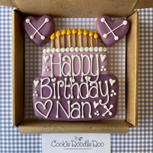 May include: Purple and yellow decorated sugar cookie in the shape of a birthday cake with the text "Happy Birthday: Nan: x" and two purple heart-shaped cookies with an X on them.