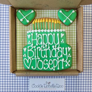 May include: A green and white decorated cookie in the shape of a birthday cake with the words "Happy Birthday Joseph" on it. The cookie is in a box with two green heart-shaped cookies.