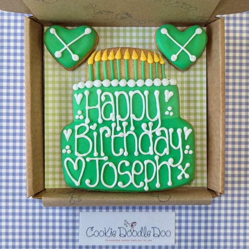 May include: A green and white decorated cookie in the shape of a birthday cake with the words "Happy Birthday Joseph" on it. The cookie is in a box with two green heart-shaped cookies.