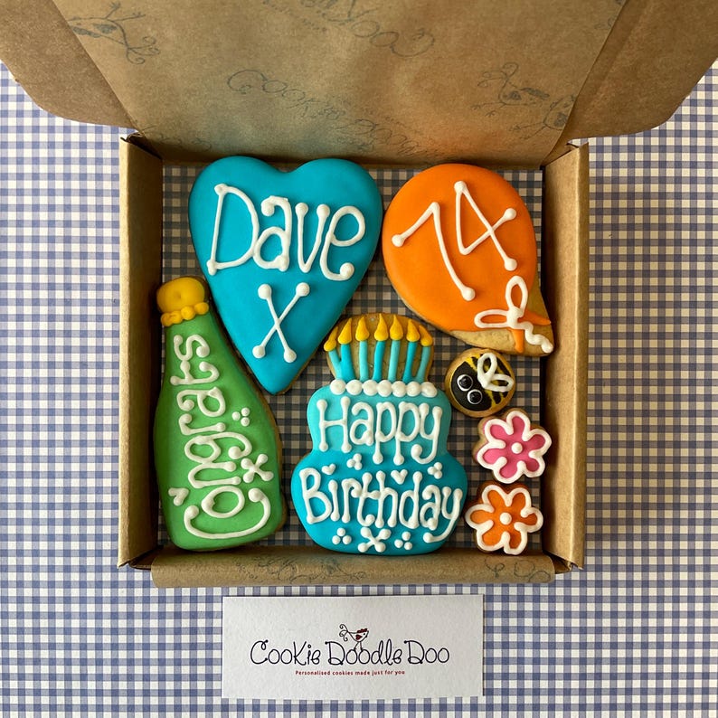 May include: A box of decorated sugar cookies with a blue heart cookie that says "Dave X", an orange balloon cookie with the number "14", a green bottle cookie that says "Congrats", a blue and white cake cookie that says "Happy Birthday", a black and yellow bee cookie, and two pink and orange flower cookies.