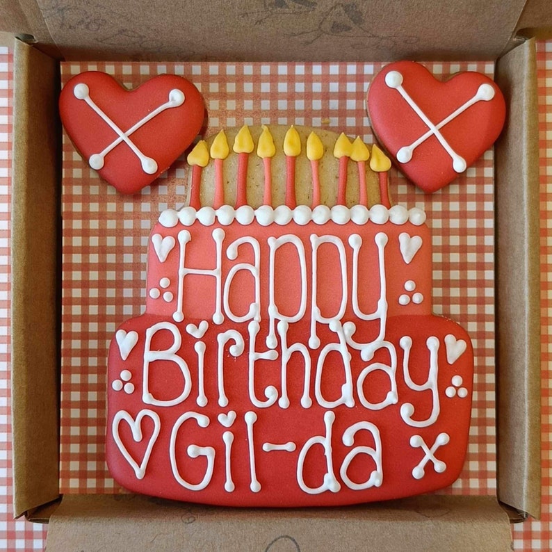 May include: A red and white decorated gingerbread cake with the words "Happy Birthday Gil-da" in white icing. The cake is surrounded by two red heart-shaped cookies with white crosses.