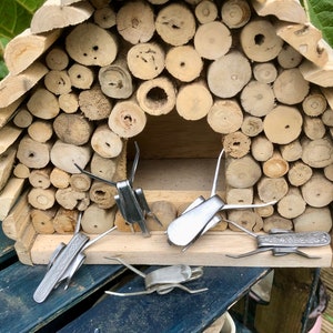 May include: Four silver metal insects are arranged in front of a wooden insect house made of stacked logs. The insects are made from recycled materials and have a rustic, handmade look.