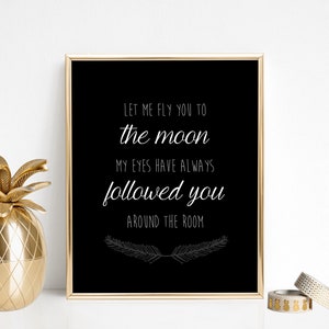May include: A framed black and gold print with the text "Let me fly you to the moon, my eyes have always followed you around the room." A gold pineapple decor and a geometric candle are also visible.