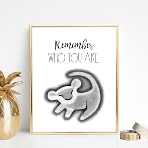 Peut inclure: Impression encadrée avec le texte « Remember WHO YOU ARE » au-dessus d'une illustration en niveaux de gris d'un lionceau. Un récipient doré en forme d'ananas et une bougie géométrique sont sur la table.