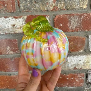 May include: A colorful glass pumpkin with a swirl design in shades of pink, yellow, blue, and white. The pumpkin has a green glass stem.