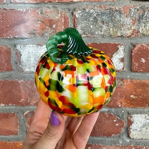 May include: A colorful glass pumpkin with a green swirled stem. The pumpkin is a mix of orange, yellow, red, and green glass pieces. The decorative fall-themed item is set against a brick background.