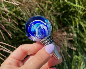 May include: A blue and white glass bottle stopper with a silver metal base. The glass is swirled with white and blue colors.