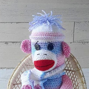 Op de afbeelding: Een gehaakte sok-aap die een roze en blauwe gestreepte muts en sjaal draagt, zittend op een rieten stoel. De aap is gemaakt van roze, blauwe en witte draad. Heartmade de Specials4u staat op de vloer.
