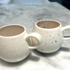 May include: Two round, off-white ceramic mugs featuring a speckled pattern with small, multi-colored dots in shades of blue, orange, yellow, and green. Each mug has a smooth, glossy finish, a wide, rounded body, and a single curved handle on the side.