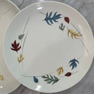 May include: Two off-white ceramic plates with a speckled finish. The plates are decorated with a leaf design in shades of blue, yellow, and brown. One plate is oval, and the other is round. The plates are likely for serving food.