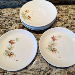 May include: A set of white ceramic plates and saucers. Each piece features a delicate design of autumn leaves in shades of brown, green, and yellow. The plates are round and oval-shaped, perfect for serving meals.