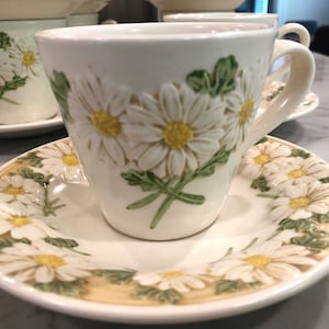 May include: White teacup and saucer set with embossed daisy floral design. The teacup features a handle and is placed on a matching saucer. The daisy design includes white petals, yellow centers, and green leaves. The set is made of ceramic.