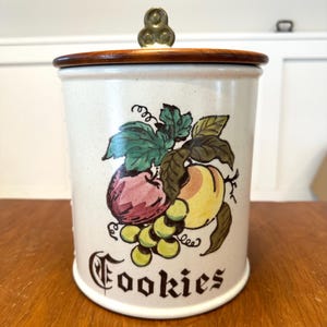 May include: Cream-colored ceramic cookie jar with a wooden lid and brass handle. The jar features a colorful illustration of fruit, including grapes, peaches, and plums, with the word "Cookies" in a decorative font.