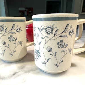 May include: Set of ceramic mugs with a speckled cream background and a blue floral design. Each mug has a square handle and a blue stripe around the rim. The mugs are decorated with a delicate floral pattern.