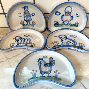 May include: A set of five crescent-shaped ceramic dishes with a white glaze and blue trim. Each dish features a hand-painted blue and orange illustration of a different subject: a person, a duck, a sheep, a dog, and a rooster, all on green grass.
