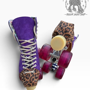 May include: A pair of roller skates featuring purple suede boots with leopard print toe caps and heel accents. The skates have beige laces with silver barbed wire charms, translucent pink wheels, and a beige toe stop. The logo reads Black Sheep Roller Skate Shop.