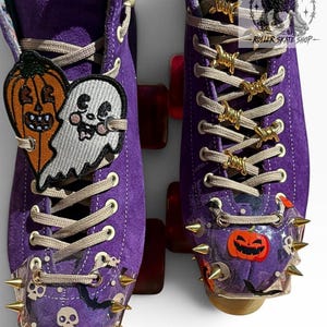 May include: A pair of purple suede roller skates featuring beige laces, gold-tone barbed wire lace charms, and clear toe guards with gold spikes. The skates are decorated with Halloween-themed patches of a pumpkin and ghost, and skull decals.