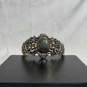 May include: A silver and gold bracelet with an oval-shaped green gemstone set in the center. The bracelet has a detailed, ornate design.