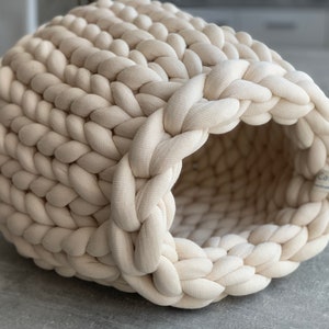 Könnte beinhalten: Ein cremefarbenes gestricktes Katzenbett mit einer runden Öffnung. Das Bett ist aus dickem, grobem Garn gefertigt und hat eine weiche, kuschelige Textur.