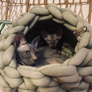 May include: A sage green, chunky knit, round pet bed with a thick braided texture. Two hairless cats are resting inside the cave-like structure. A small brown leather tag on the side features the text "MADE BY Cats&DogsDE" with a metal rivet.